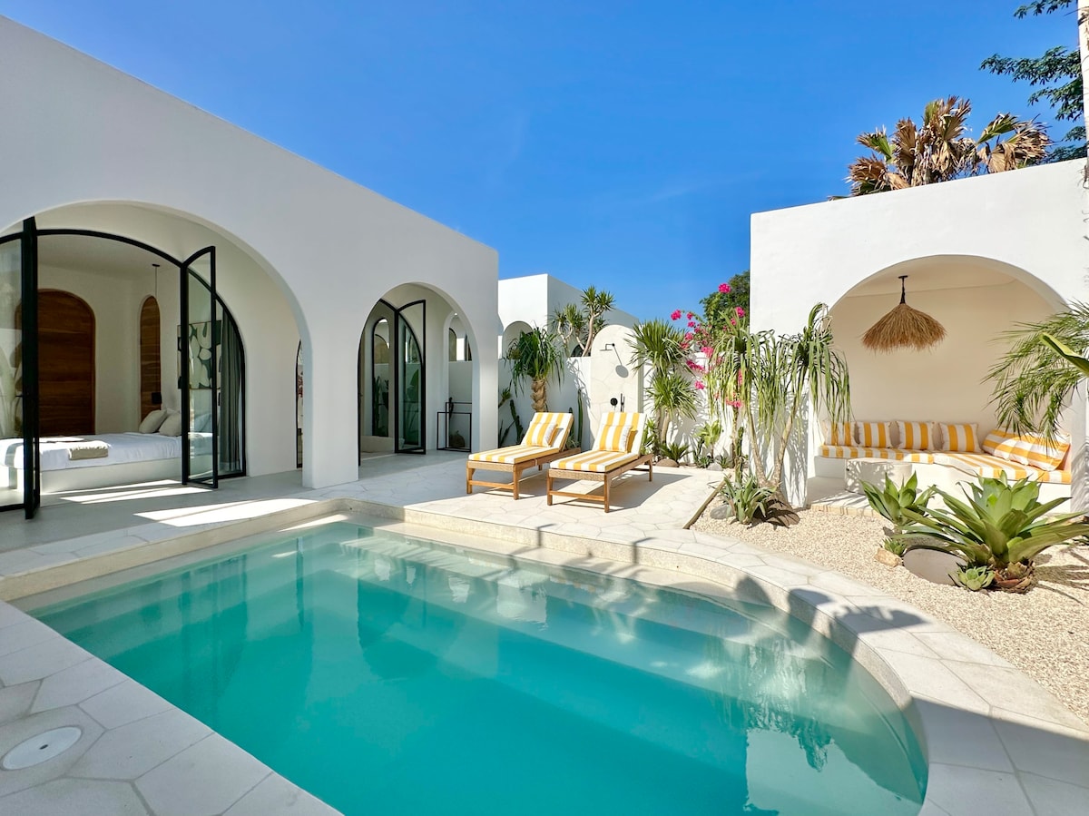 An inviting outdoor space features a shimmering private pool surrounded by tropical plants and comfortable lounge chairs. Two arched structures encompass the pool area, with one side showcasing a cozy seating arrangement and the other leading to a bright sleeping area.
