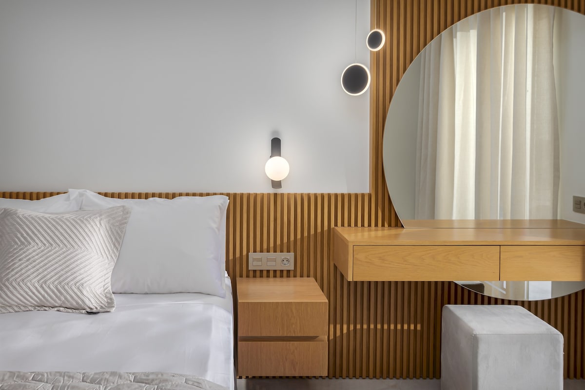 A serene bedroom area features a spacious bed with crisp linens and decorative pillows. A sleek wooden nightstand and a modern dresser with a round mirror are present. Soft, natural light filters through sheer curtains, creating a calming ambiance.