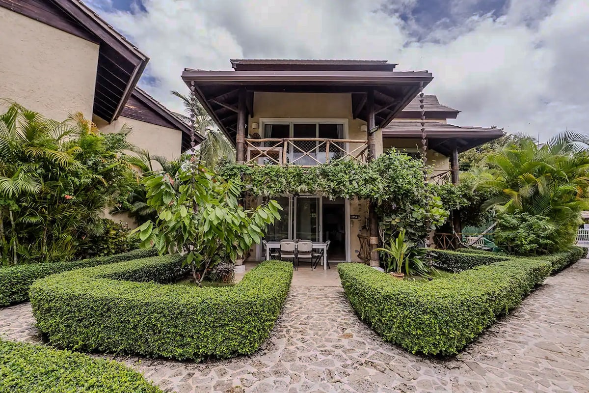The exterior of the townhouse is surrounded by well-manicured hedges and tropical plants. A stone pathway leads to a covered porch with a casual seating area, framed by wooden beams and large windows, under an overcast sky.