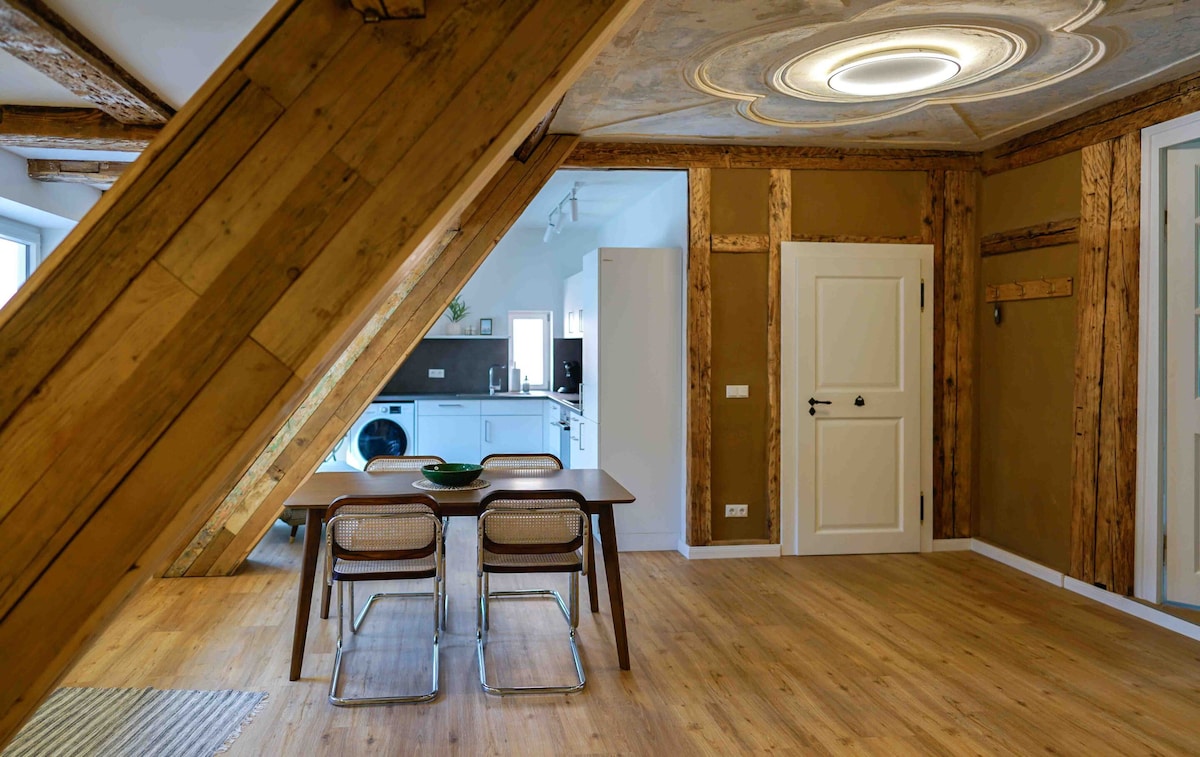 The open-plan living area features wooden beams and a decorative stucco ceiling. A dining table with four chairs is positioned in the space, which leads to a fully equipped kitchen visible in the background. Warm, earthy tones create a welcoming atmosphere.