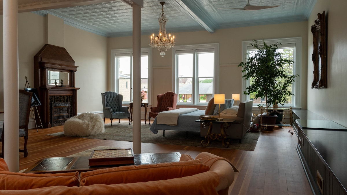 An open studio space is presented with large windows allowing natural light to fill the room. Elegant furnishings, including vintage armchairs and a spacious bed, are arranged around a central seating area. A decorative chandelier hangs above, with a plush area rug adding warmth to the hardwood floor.