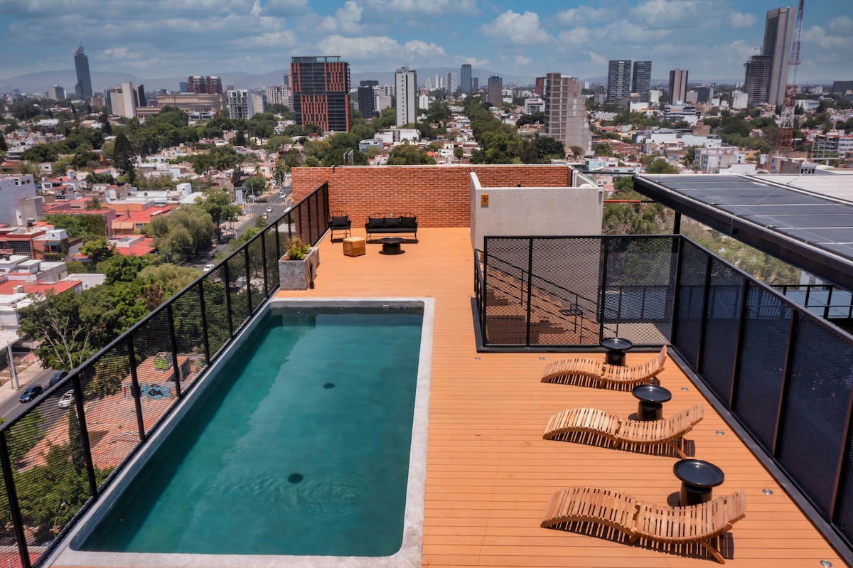 A rooftop area features a refreshing pool surrounded by wooden decking. Sun loungers are placed along the edge, offering a view of the city skyline. The vibrant cityscape is visible in the background, creating a serene outdoor space for relaxation.