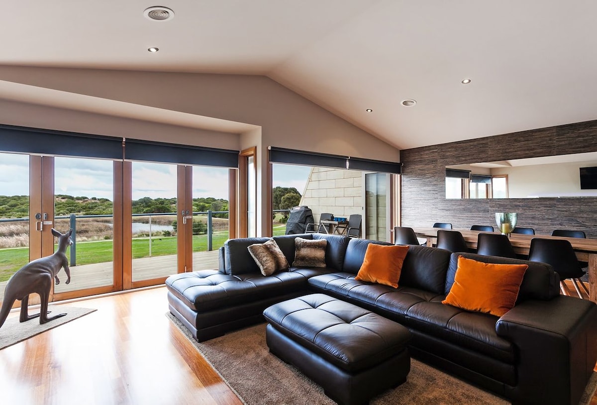 A spacious lounge area features a dark leather sectional sofa adorned with orange cushions. Bi-fold doors open to an outdoor deck, offering views of the coastal landscape. Natural light floods the room, which includes a dining table and stylish decor.
