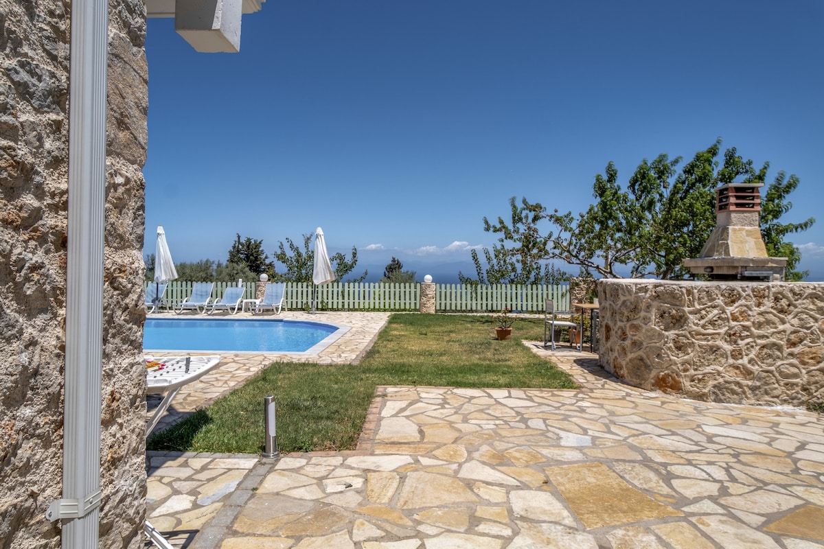 A spacious outdoor area features a swimming pool surrounded by sun loungers. A stone terrace leads to a well-maintained lawn, with trees providing shade. The view encompasses the Ionian Sea, and a barbecue station is visible nearby, enhancing the outdoor experience.