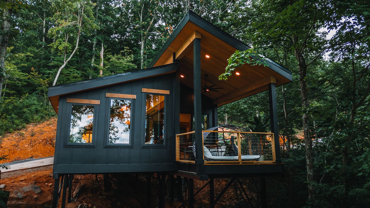 The treehouse is elevated among lush greenery, featuring a modern exterior with dark wood siding. Large windows enhance the connection to the outdoors, while a covered deck showcases comfortable seating, inviting relaxation in a serene natural setting.