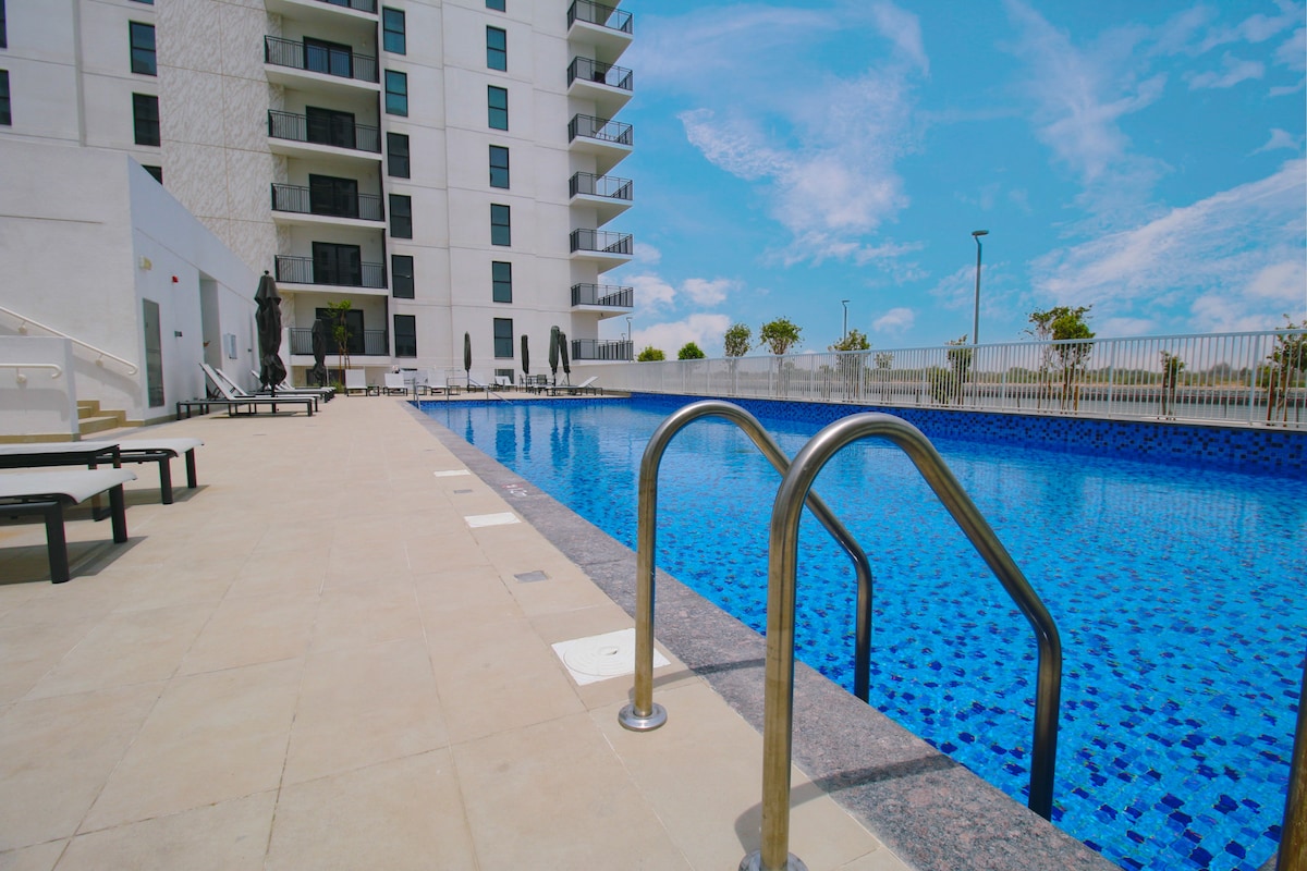 A large swimming pool glistens under a clear blue sky, bordered by light-colored stone tiles. Lounge chairs line the poolside, while a few black umbrellas provide shade. The building's modern architecture is visible in the background, emphasizing the spacious outdoor area.
