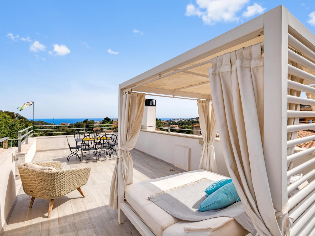 An outdoor terrace features a sunbed draped with light curtains and accented by colorful cushions, providing a serene spot for relaxation. A dining area with a round table and chairs overlooks the landscape, enhanced by views of the nearby ocean and greenery in the distance.