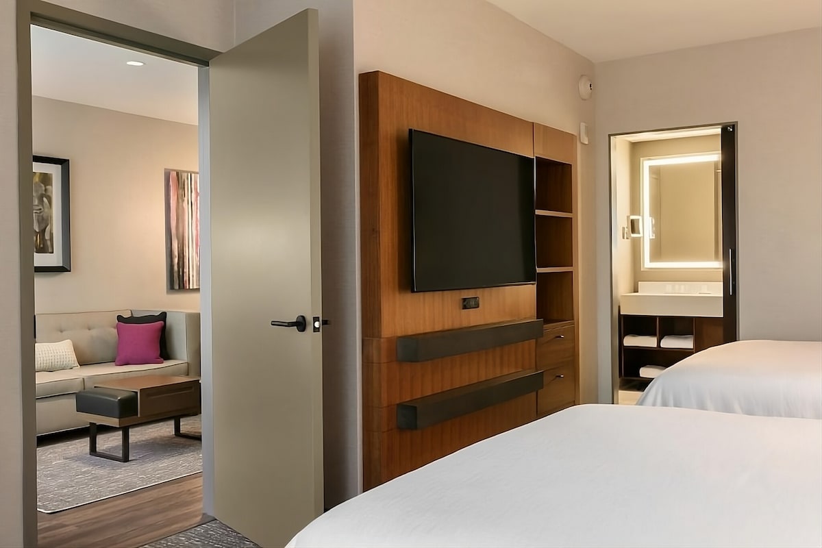 The spacious hotel suite features two queen beds and a seating area with a sofa adjacent to a coffee table. A wall-mounted flat-screen TV is visible along with a doorway leading to a bathroom area with vanity lighting.
