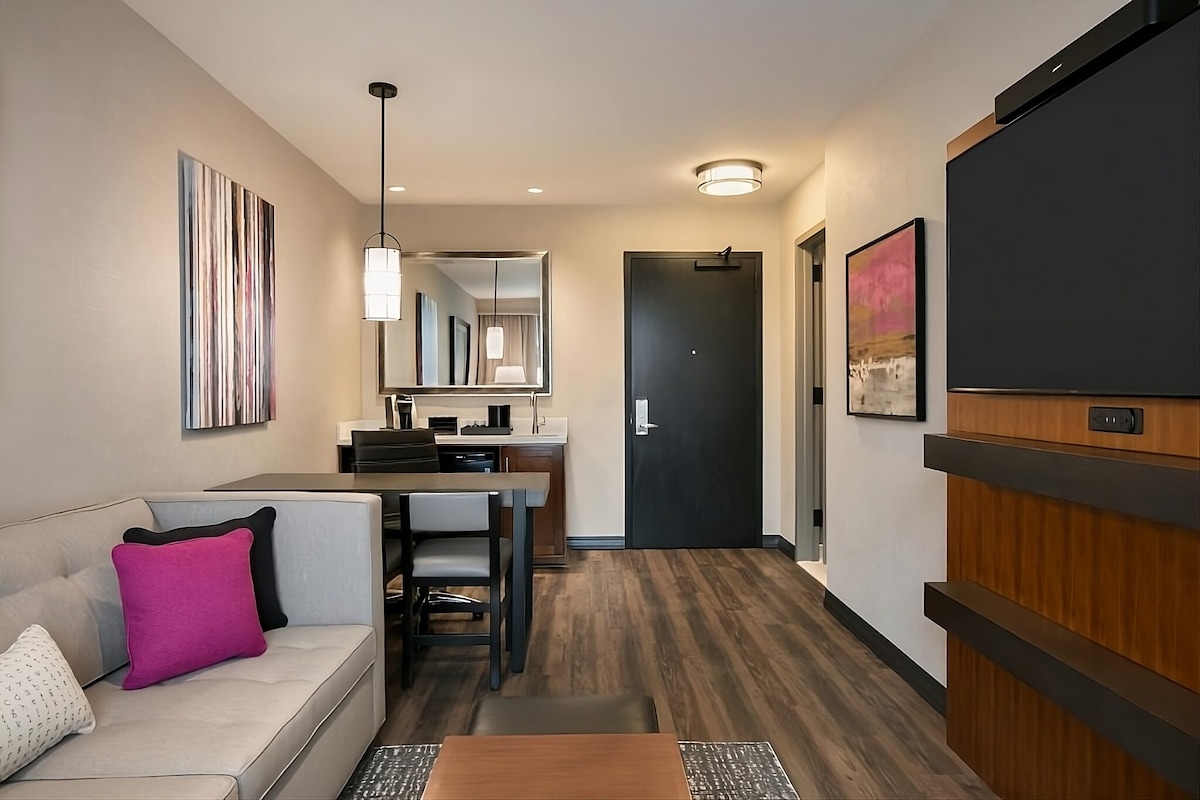 A modern hotel suite is presented with a comfortable seating area featuring a light-colored sofa and a dining table. A flat-screen TV is mounted on the wall, and artwork decorates the space, adding subtle color. The entrance door and additional furnishings are visible, creating a welcoming atmosphere.