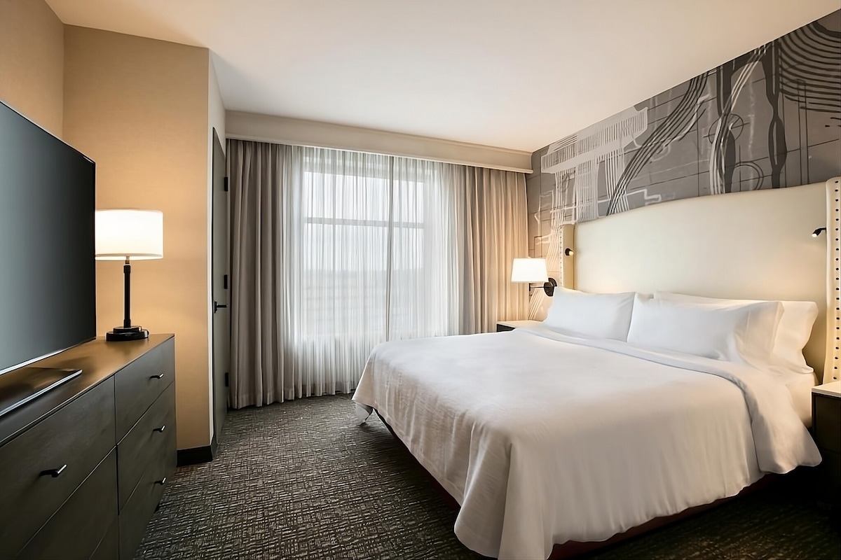 A spacious hotel room is designed for comfort, featuring a king bed with crisp white linens. A large flat-screen TV is positioned across from the bed, and natural light filters through sheer curtains, enhancing the inviting atmosphere. A bedside lamp provides additional lighting.