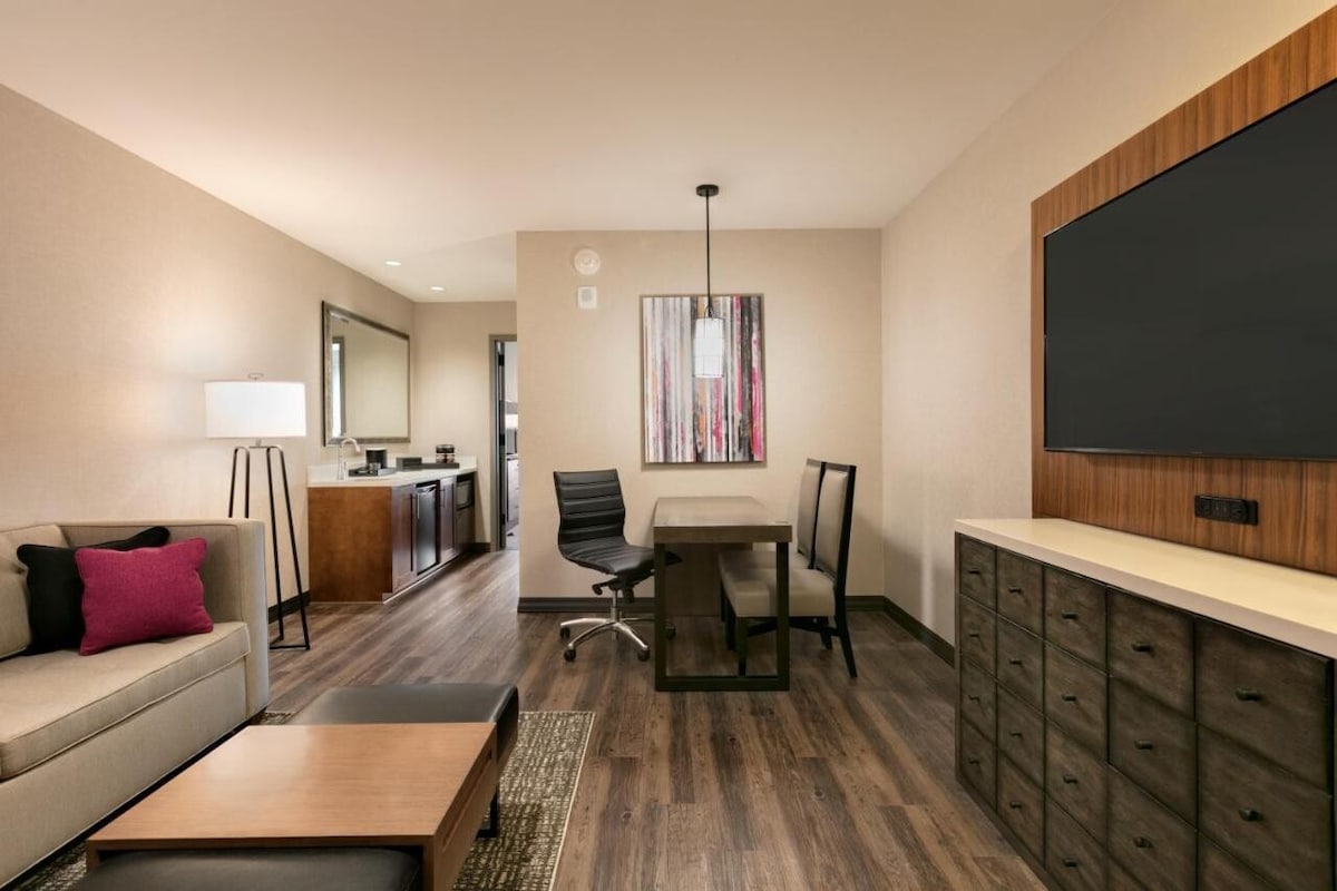 The spacious suite features a seating area with a sofa and a coffee table. A dining table with black chairs is set against light-colored walls, complemented by wooden cabinetry. A large flat-screen TV is mounted on the wall, providing a contemporary touch to the welcoming space.
