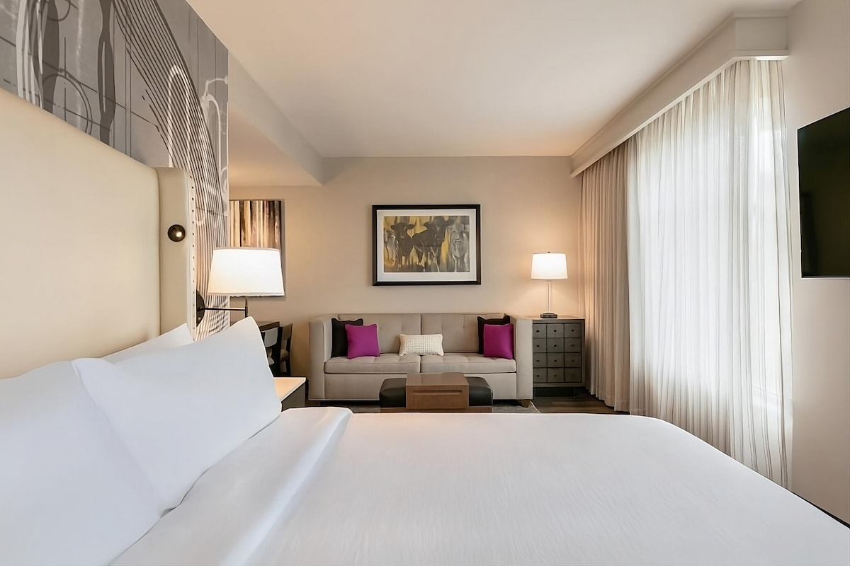 A spacious hotel room features a comfortable king bed with crisp white linens. A seating area contains a sofa and colorful accent pillows. Large windows are dressed with sheer curtains, allowing natural light to fill the room. A flat-screen TV is mounted on the wall.