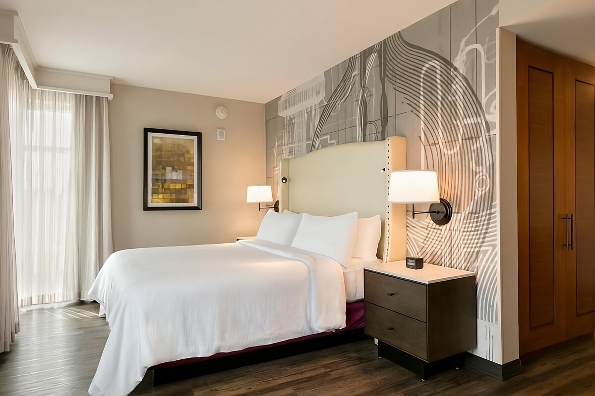 A spacious hotel room features a king bed with white linens, complemented by a neutral headboard. A contemporary art piece is displayed on the wall, alongside a light wooden wardrobe. Soft natural light filters through sheer curtains, enhancing the modern decor.