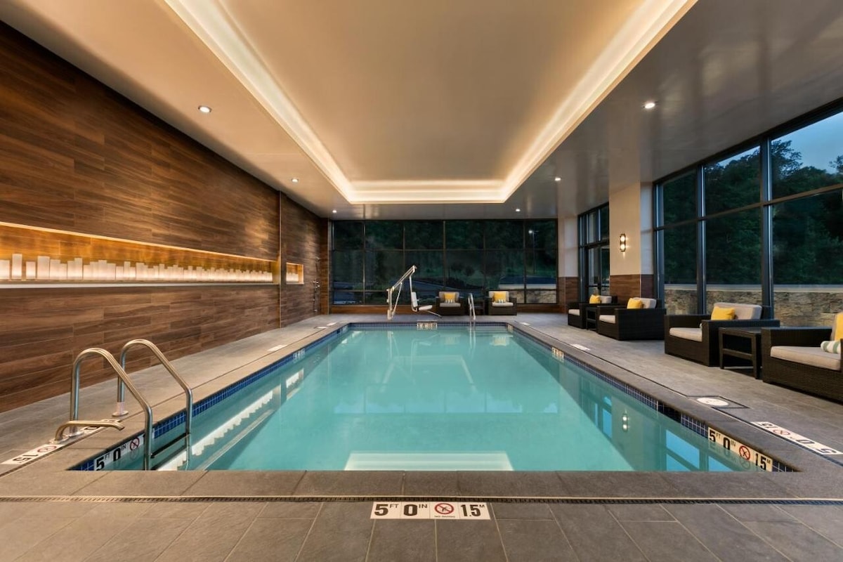 An indoor swimming pool area is illuminated with soft lighting, featuring sleek tile flooring and a wooden accent wall. Comfortable seating is arranged along the poolside, with large windows offering a view of the outdoors. Depth markings are visible along the pool's edge.