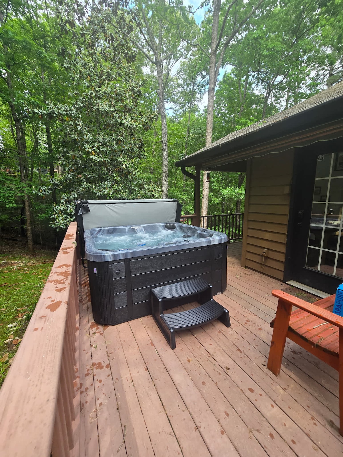 A hot tub is positioned on a wooden deck surrounded by lush greenery. A short set of steps leads up to the tub, which is partially covered with a lid to maintain temperature. The adjacent wooden seating provides a spot to relax amidst nature.