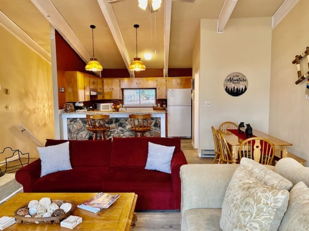A bright living area is visible, featuring a red sofa and a wooden coffee table with decorative accents. The open-concept layout includes a dining area with wooden chairs and a well-equipped kitchen in the background, illuminated by pendant lighting.