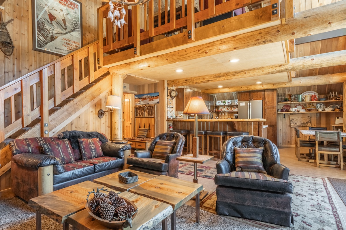 The spacious living area features a cozy seating arrangement with a dark leather sofa and two matching armchairs, complemented by a wooden coffee table. Light brown wooden beams and walls add a natural feel, while a staircase leads to the lofted area above. Subtle lighting creates a warm ambiance.