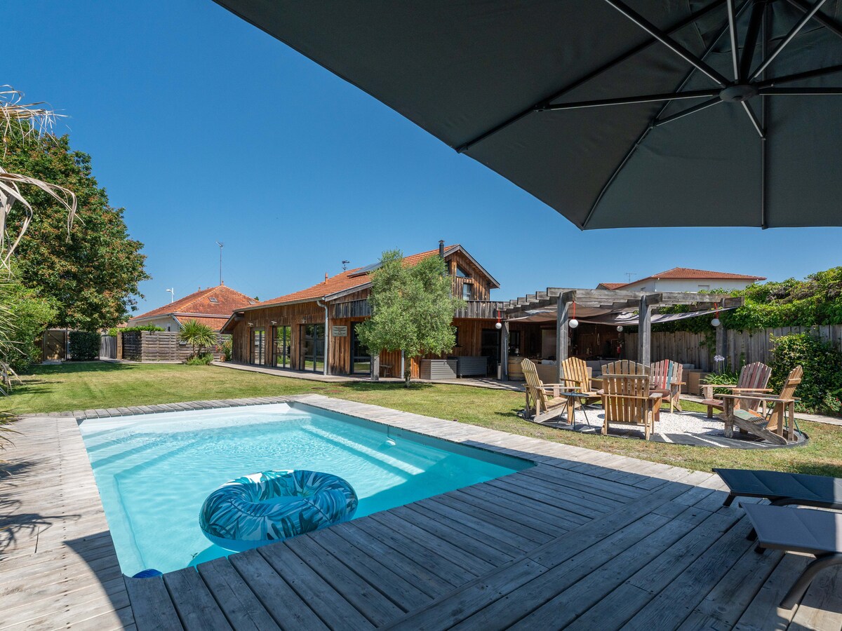 A serene outdoor space features a heated pool surrounded by a wooden deck. A spacious garden is visible with leisure seating, complemented by an expansive property in the background. Clear skies provide ample sunlight, enhancing the inviting atmosphere of the relaxation area.