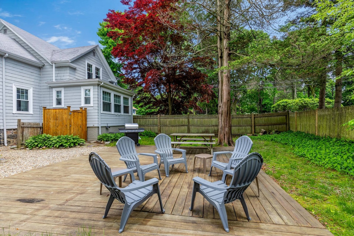 An outdoor deck area features a round table at the center, surrounded by six Adirondack chairs. Lush green grass and various trees provide a serene backdrop, while a picnic table is positioned nearby. A gas grill is visible against a wooden fence.
