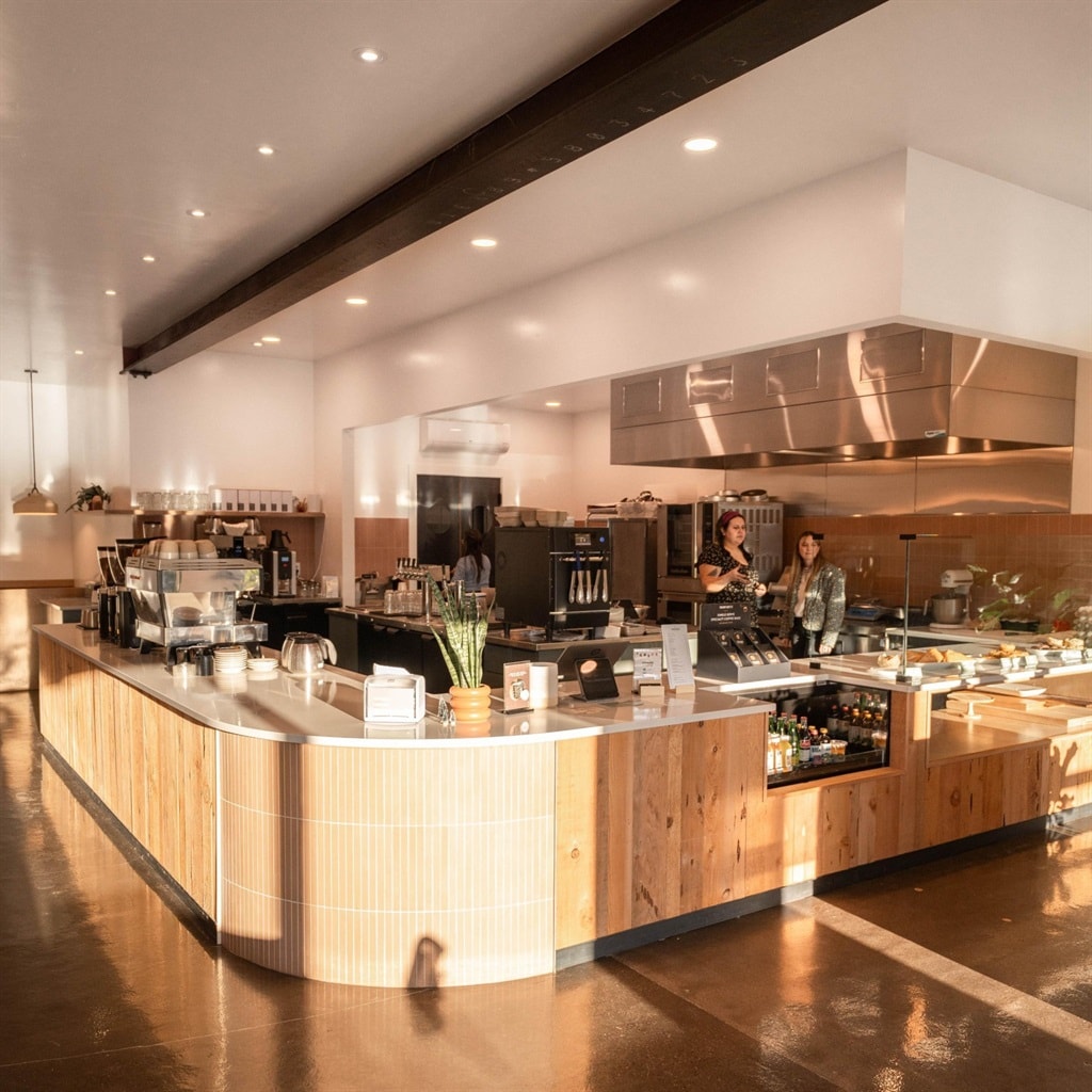 A welcoming café interior features a spacious wooden counter with a variety of food and beverage options. Warm lighting enhances the modern design, while an open kitchen allows views of chefs preparing meals. Natural elements, such as potted plants, are incorporated throughout the space.