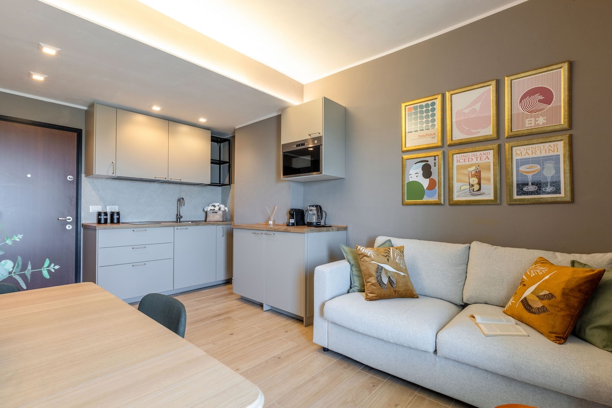 A well-designed living area features a light grey sofa adorned with decorative pillows, accompanied by a wooden dining table. The fully equipped kitchen is visible, with modern appliances and cabinetry, complemented by framed artwork on the wall.
