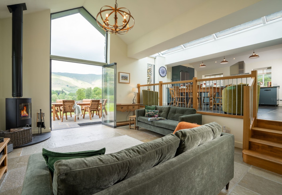 The spacious open-plan living area features a green sofa arranged around a wood-burning stove. A large glass door invites natural light and offers views of the outdoor dining area and surrounding hills. The vaulted ceiling enhances the room’s airy feel.