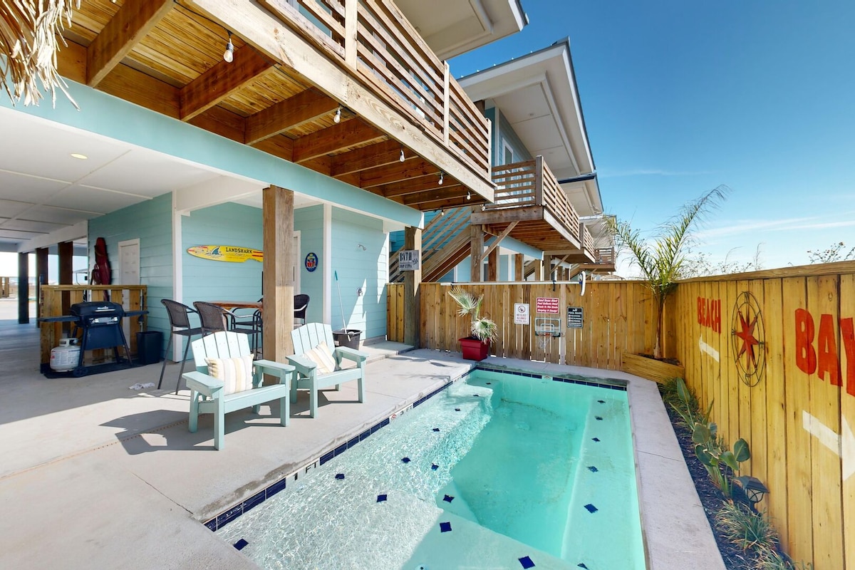An outdoor area features a private pool surrounded by a concrete deck with lounge chairs. Nearby, a propane grill is placed next to a vibrant wooden fence adorned with coastal decor. The space is illuminated by soft lighting, enhancing the inviting outdoor atmosphere.