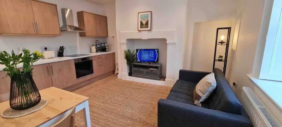 A modern kitchen area features light wood cabinetry and stainless steel appliances. A comfortable sofa sits beside a television in an inviting living space, illuminated by natural light from a window. A simple table is set for dining, accompanied by a vase of fresh flowers.