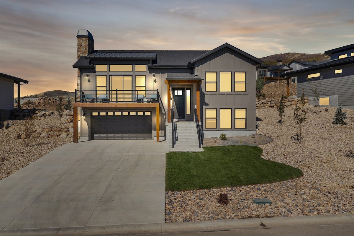 A modern home is presented with a stone and wooden facade, showcasing large windows that reflect the natural surroundings. A spacious driveway leads to a two-car garage, while landscaped areas feature gravel and minimal greenery for easy maintenance.