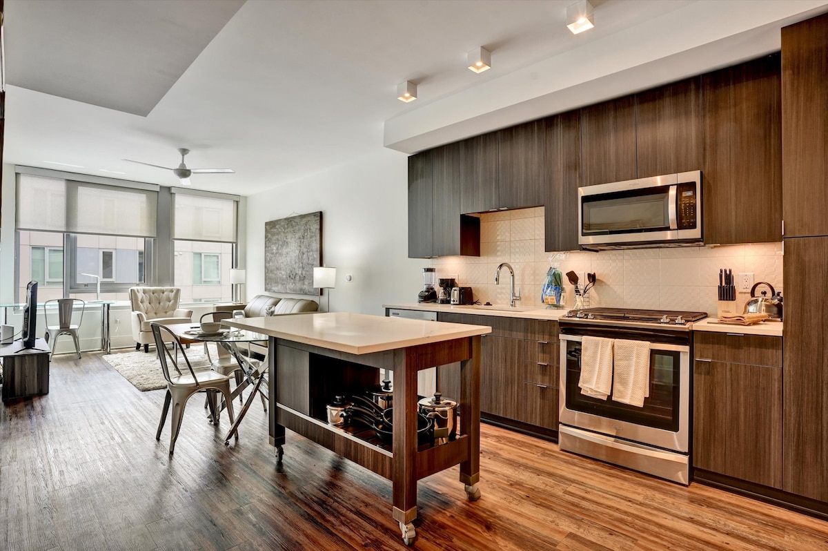 A modern kitchen with stainless steel appliances is featured alongside a spacious island. Natural light floods the area through large windows, illuminating the sleek wooden cabinets. An inviting living space with a comfortable armchair and dining table is situated adjacent to the kitchen.