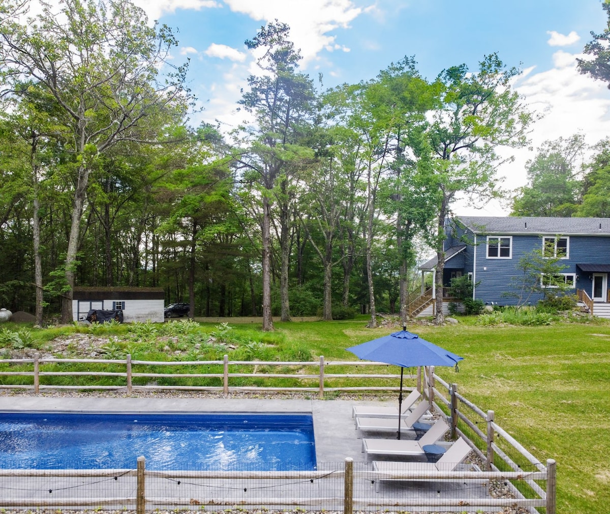 A refreshing swimming pool is surrounded by a spacious patio with lounge chairs and an umbrella for shade. Mature trees create a serene backdrop, while a blue house and a firepit area are visible in the distance, enhancing the natural ambience.