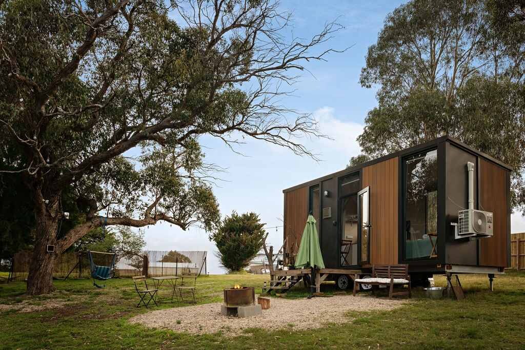 A tiny home with a contemporary design is set among lush greenery, featuring large windows that open to the outdoors. A cozy fire pit area is surrounded by outdoor seating, while a large eucalyptus tree provides shade nearby, enhancing the connection with nature.