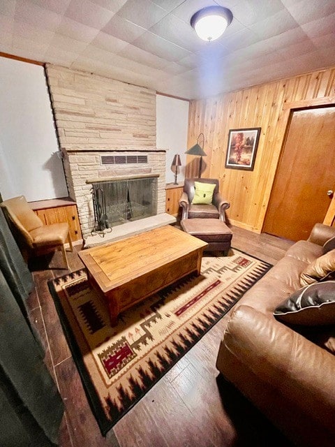 A cozy living area features a stone fireplace as its focal point, surrounded by wooden paneling. A wooden coffee table rests on a patterned rug, with a comfortable armchair and a leather sofa providing seating options. Natural light is diffused through curtains on one window.