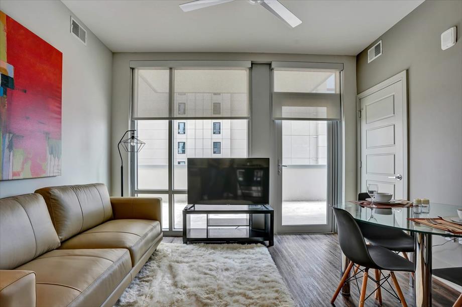 The living area features a comfortable tan sofa paired with a contemporary coffee table and a sleek television stand. Large windows provide natural light, and a dining set with black chairs adds a modern touch. A plush area rug enhances the space.