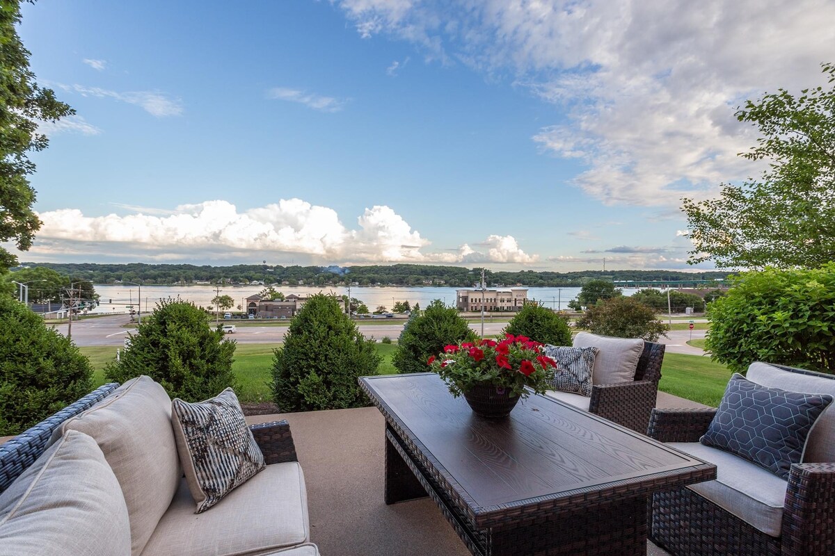 A comfortable seating area features plush couches and a wooden coffee table, surrounded by lush greenery. The view overlooks the Mississippi River with a sky filled with clouds, offering a serene outdoor space for relaxation.