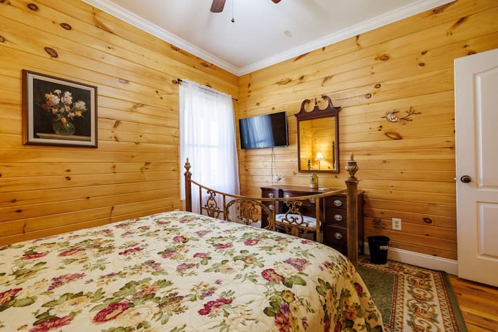 საძინებელი, რომელშიც დგას საწოლი 152×203 სმ ზომის ლეიბით, ხის თბილი კედლები, ვინტაჟური რკინის საწოლი ყვავილებიანი თეთრეულით, კომფორტული გარემო, კედელზე დამონტაჟებული ტელევიზორი და ანტიკვარული ტუალეტის მაგიდა — იდეალურია დასასვენებლად და ნოსტალგიისთვის.