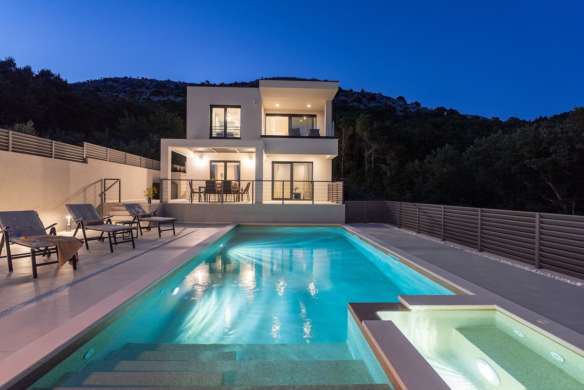 A modern villa is illuminated under the evening sky, featuring a heated swimming pool with a hydromassage area. Comfortable deck chairs surround the pool, and glass doors provide views into the main living area. Lush greenery borders the property, enhancing the serene environment.
