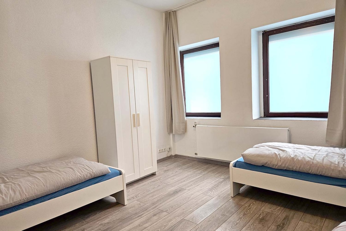 A bright room designed for comfort features two single beds with light bedding. Natural light is allowed in through two windows, with sheer curtains softly framing them. A white wardrobe stands against one wall, complemented by a neutral-colored floor.