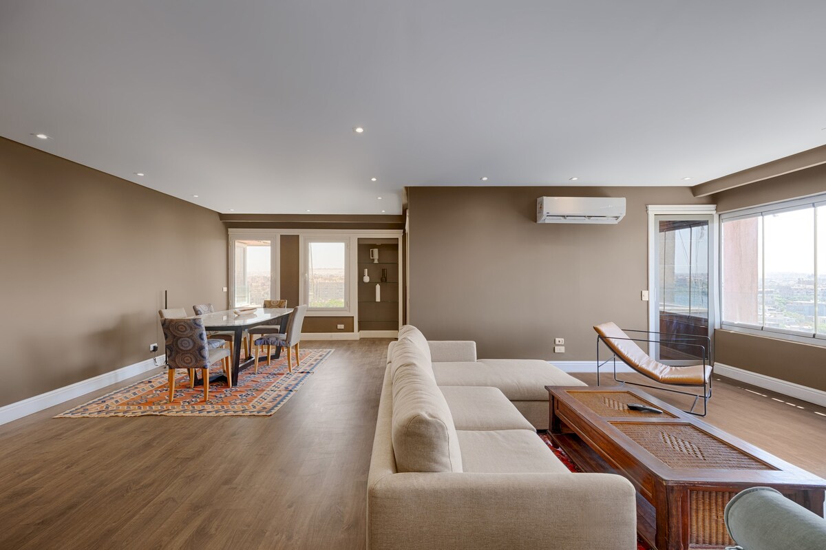 A spacious living area features neutral-colored walls and natural light from large windows. A light-colored sofa is positioned in the foreground, facing a wooden coffee table. A dining table with four chairs is visible in the background, complemented by a patterned area rug.