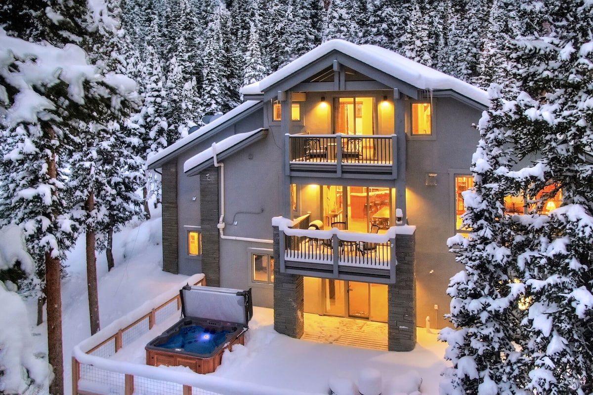 The exterior view of the chalet is surrounded by snow-covered pine trees, showcasing multiple balconies. Soft lighting emanates from the windows, highlighting the inviting atmosphere. A private hot tub is visible on the patio, providing a cozy retreat amidst the winter landscape.