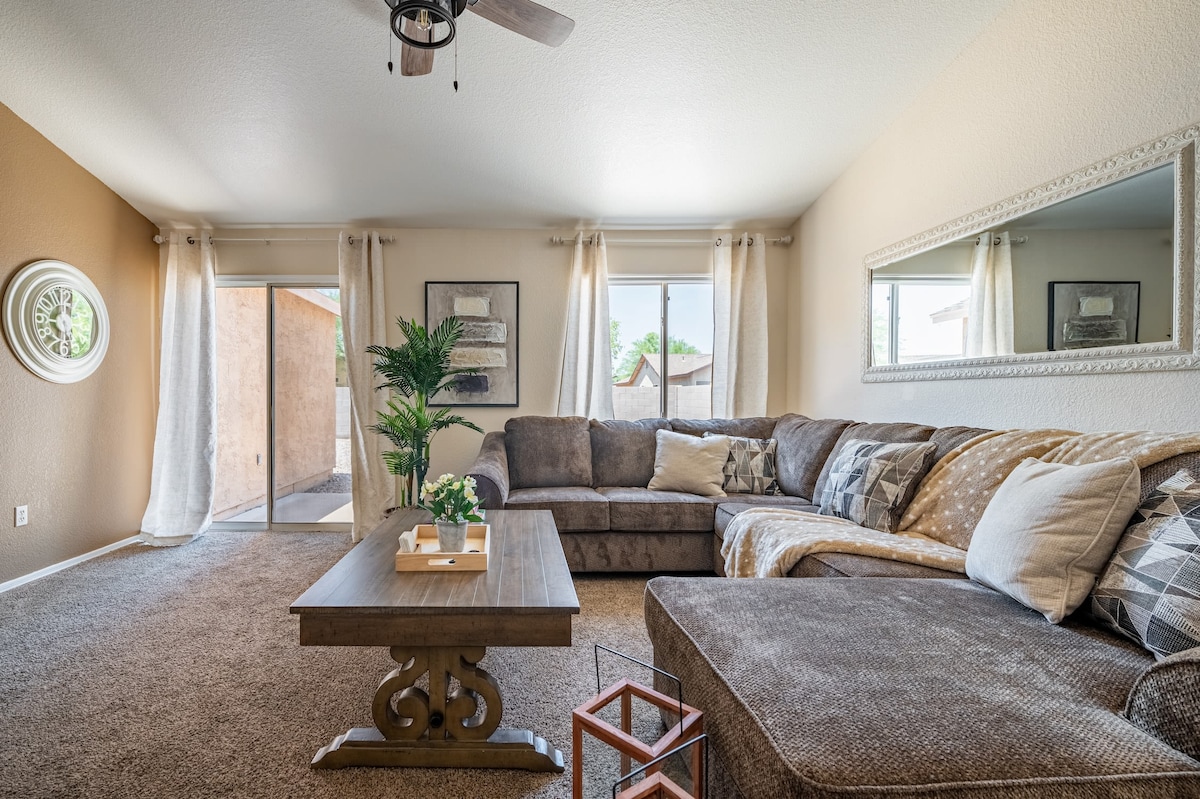 A spacious living area features a large sectional sofa adorned with decorative pillows. A coffee table rests on a plush carpet, with a small decorative item on top. Natural light streams in through two sliding glass doors adorned with sheer curtains, creating a warm and inviting ambiance.