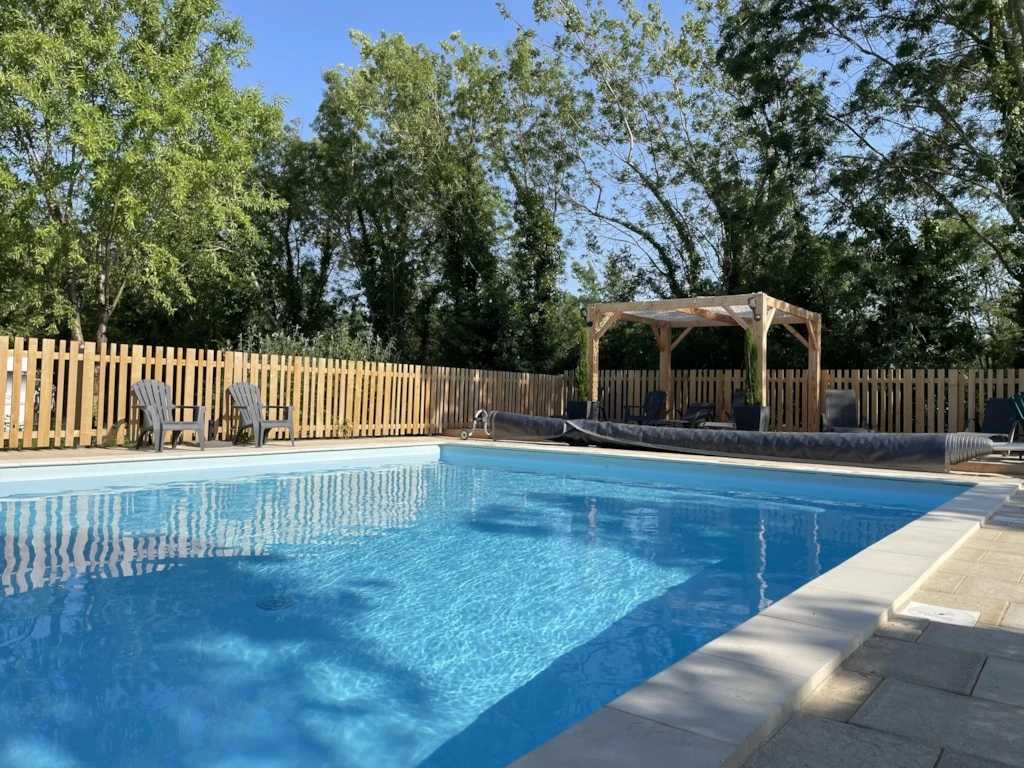 A clear swimming pool is surrounded by a wooden fence and shaded by trees. Comfortable lounge chairs are positioned nearby, offering a space for relaxation. A gazebo is visible in the background, providing additional shaded seating.