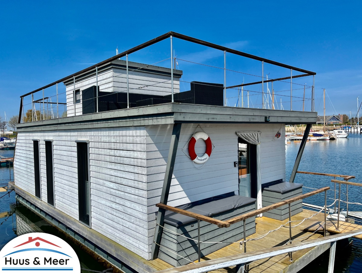 The exterior of the houseboat is presented, highlighting its modern design with a large terrace and inviting seating areas. The boat is docked at the harbor, surrounded by water and other boats, offering an unobstructed view of the sky.
