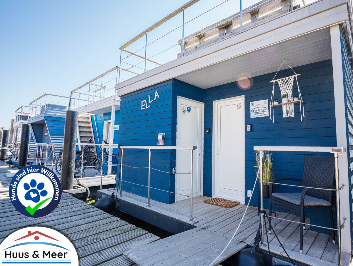 The exterior of the houseboat ELLA is presented, featuring a deep blue façade and a welcoming entrance. Wooden decking extends to the front door, while a small hanging décor piece adds character. The surroundings reflect a serene marina atmosphere.