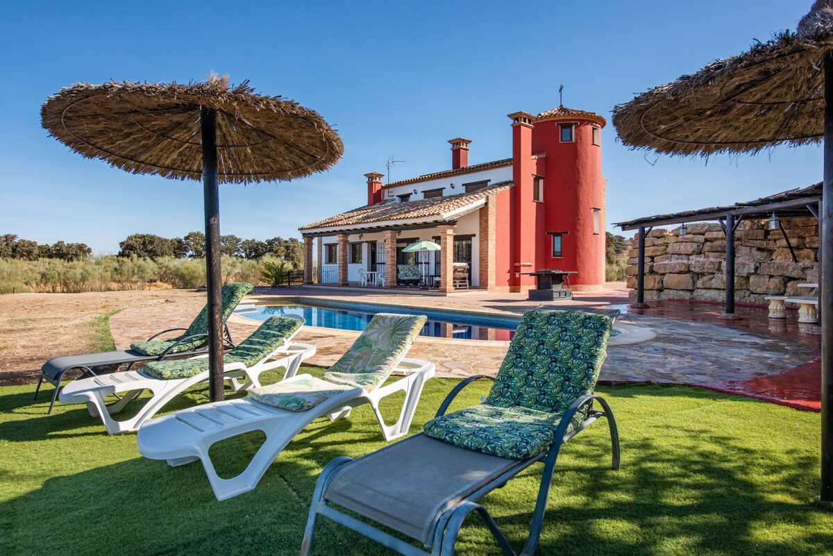 Lounge chairs with floral cushions are arranged around a private pool, under shade from thatched umbrellas. In the background, the villa is highlighted by its distinctive red tower, accompanied by a covered terrace and outdoor dining area.