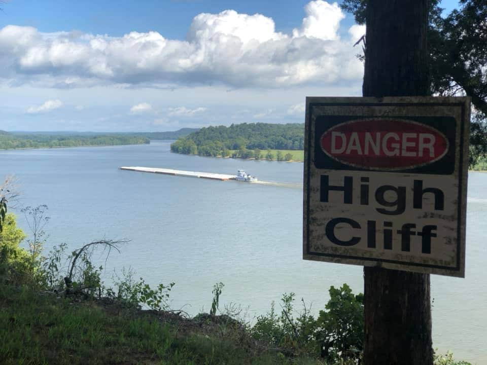 A warning sign indicating 'Danger High Cliff' stands prominently beside a scenic view of a river, bordered by lush greenery. In the distance, a barge moves along the water under a partly cloudy sky, demonstrating the area's natural landscape and waterways.