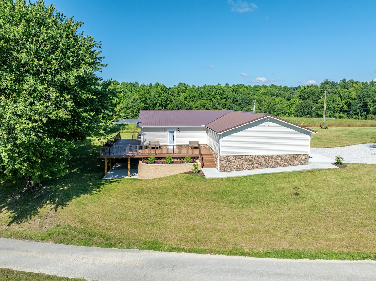 An aerial view captures a spacious single-story home surrounded by greenery. The exterior features a metal roof, stone accents, and a large deck that extends from the front. Mature trees provide shade, and a gravel driveway leads up to the property.