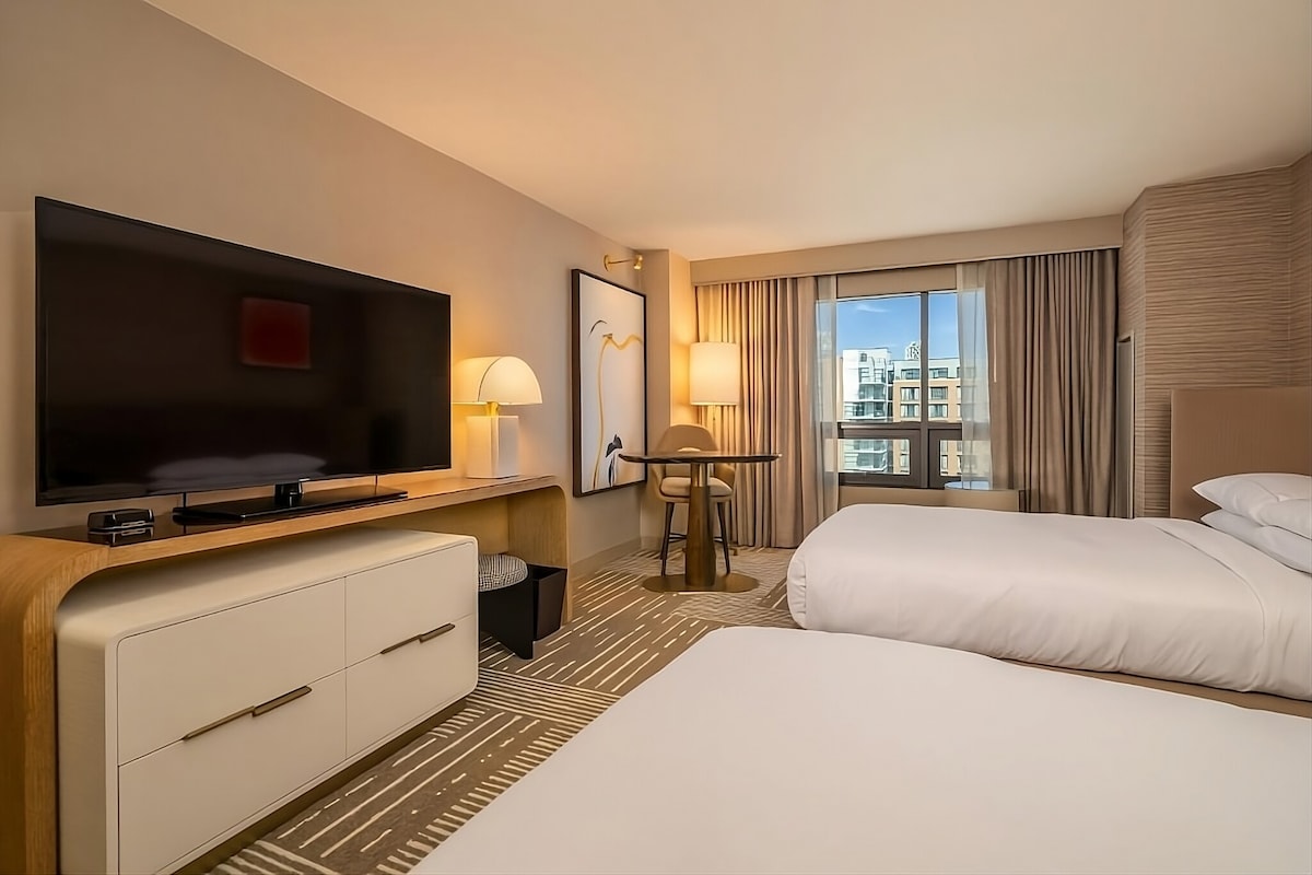 A well-appointed hotel room is visible, featuring two double beds with crisp white linens. A flat-screen TV is mounted on the wall opposite. A small round table with a lamp is positioned beside a large window covered by light drapes, providing natural light to the space.