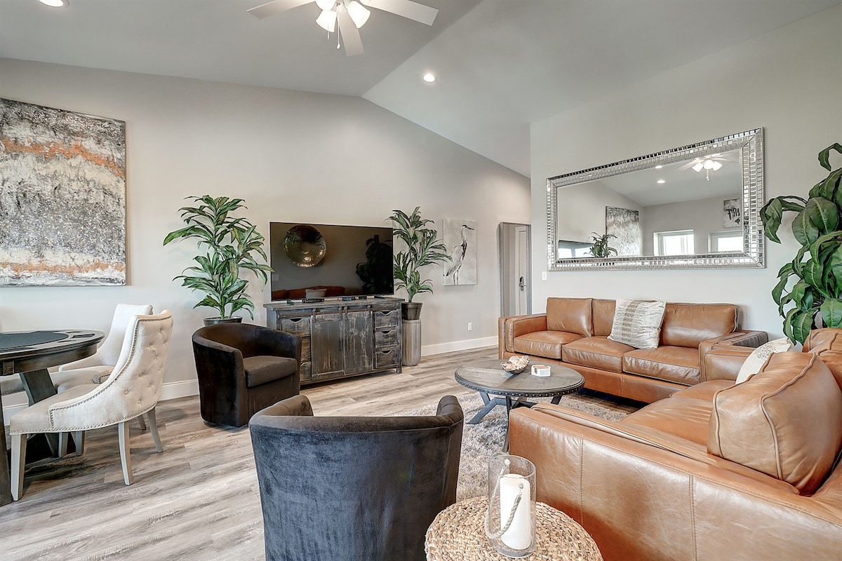 A spacious living room features stylish leather sofas and a mirrored wall, enhancing the contemporary aesthetic. A large television is mounted on the wall opposite a dining area with upholstered chairs. Potted plants provide a touch of greenery, complementing the warm color palette.