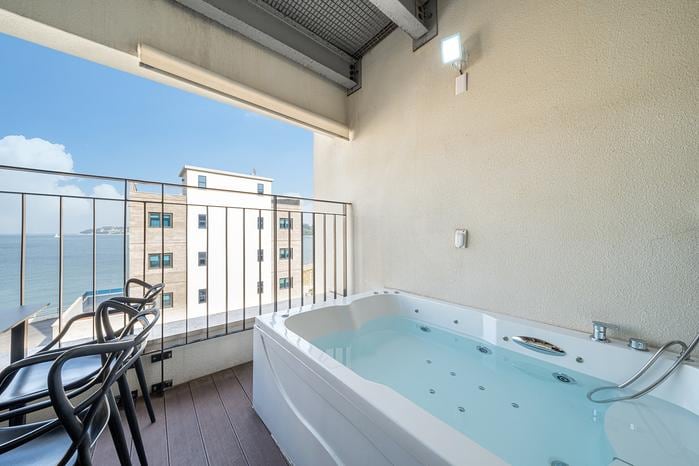 An outdoor spa is positioned on a private balcony, overlooking the ocean. The spa features a clear water surface with jets visible. Two black chairs are placed beside the spa, and a beige wall provides a backdrop, while the expansive ocean view is highlighted by clear skies.